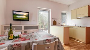 A dining area with a table set for two, a television mounted on the wall, and a kitchen with cabinets and a window. - Lavrič Garden Apartment 2 at Lake Bloke, Bloke, Slovenia (Bloke)
