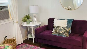 A room with a purple sofa, a round mirror, a small table with a lamp, a wicker basket, and a colorful rug.