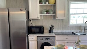 A modern kitchen with a stainless steel refrigerator, a built-in oven, a microwave, a washing machine, and a sink. There are white cabinets, a marble countertop, and a window providing natural light.