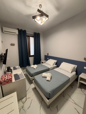 Two single beds with grey bedding and white pillows, a modern ceiling light fixture, and a small nightstand with a lamp.
