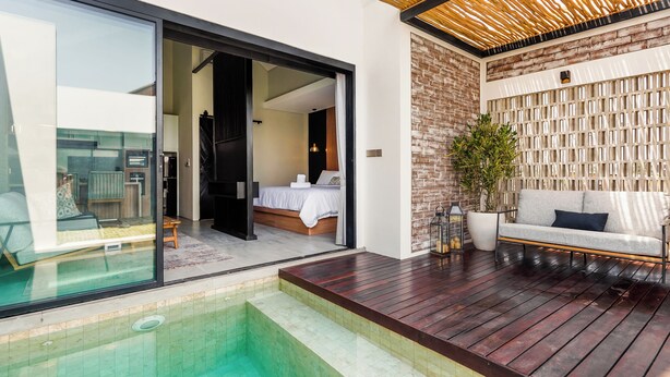 Un área de piscina moderna al aire libre con un deck de madera, una sala con paredes de vidrio que contiene una cama y una zona de estar con un sofá y una planta en maceta.