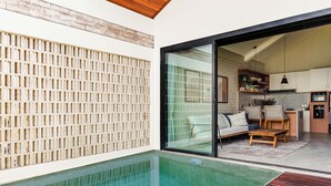 Un área de piscina moderna al aire libre con terraza de madera, cerramiento de vidrio y vista a un espacio de estar con sofá y sillones.