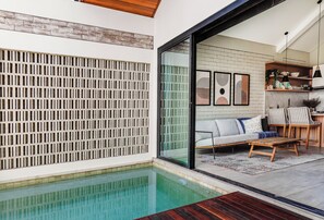 A modern pool area with a wooden deck, a white wall with a brick pattern, and a glass door leading to a living area with a sofa and chairs.
