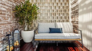 Un área de estar exterior moderna con un sofá gris, una planta grande en maceta y dos faroles.