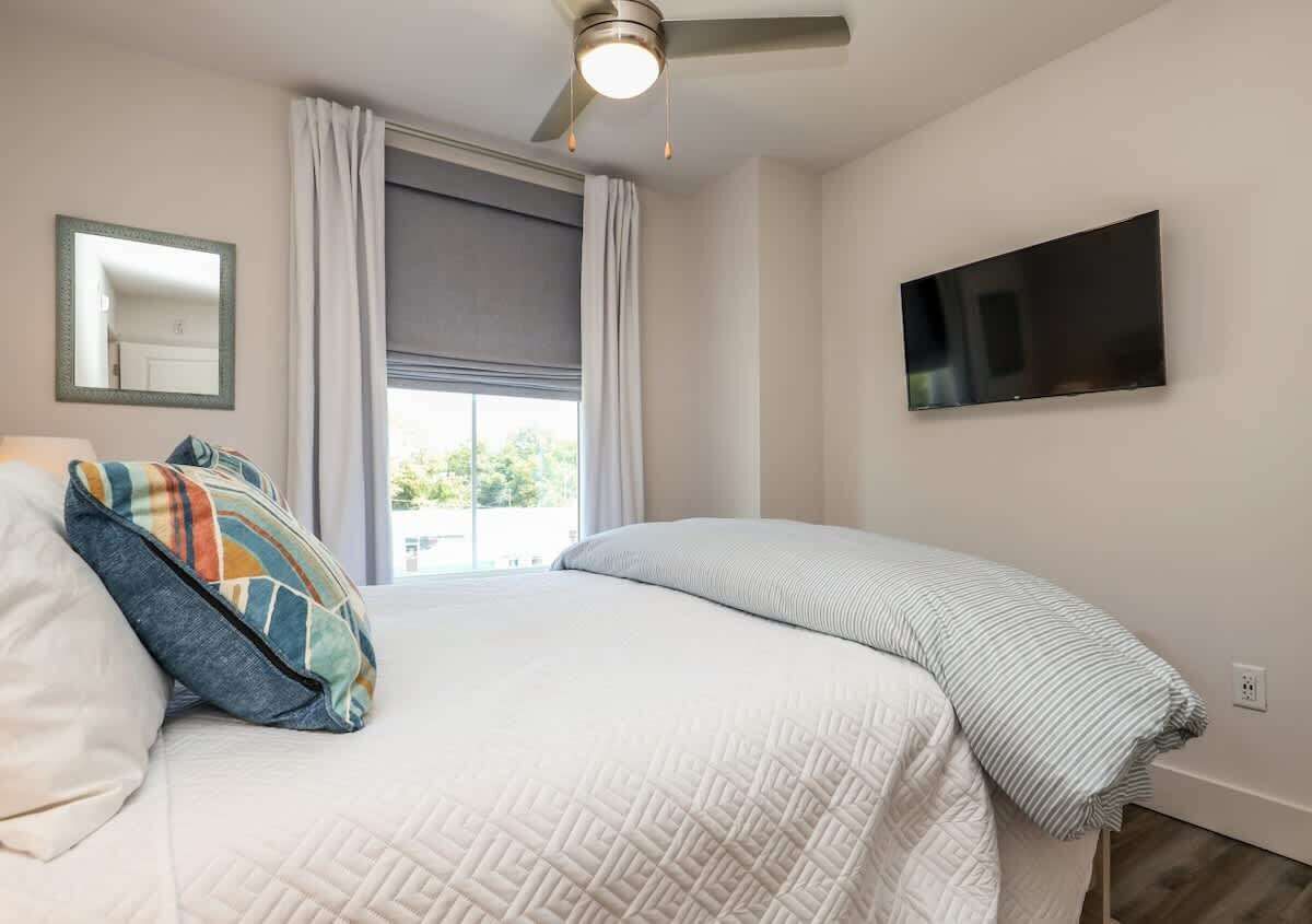 Un dormitorio con cama, ventilador de techo, televisor en la pared y ventana con cortinas.