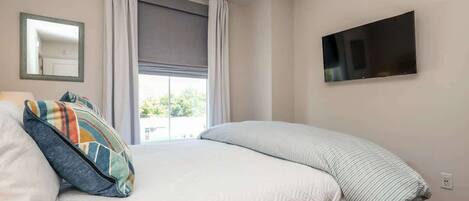 Un dormitorio con cama, ventilador de techo, televisor en la pared y ventana con cortinas.