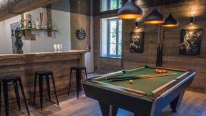 Une table de billard dans une pièce aux murs en bois, avec des tabourets de bar et un coin bar où sont exposées des bouteilles.