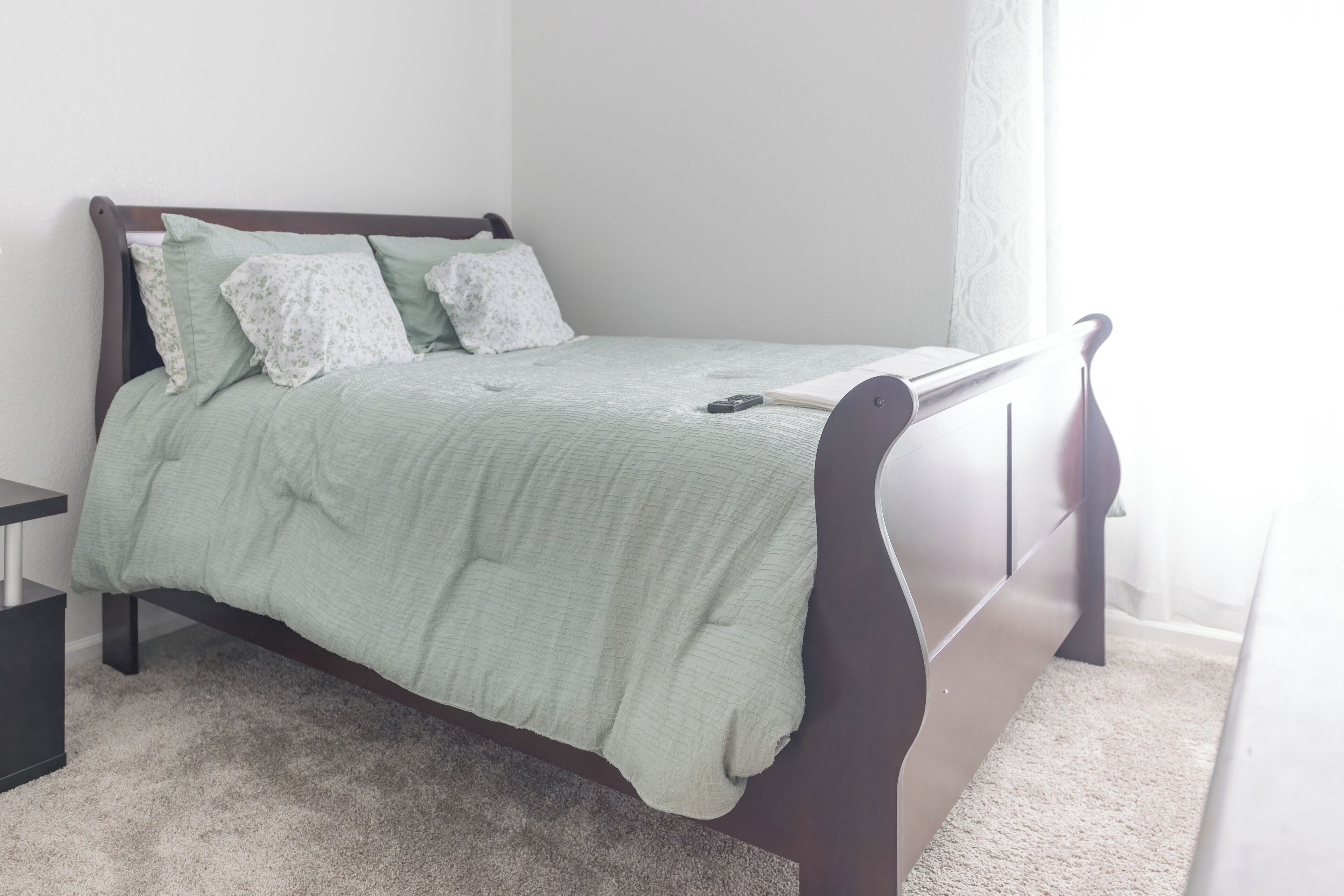 A neatly made bed with a green bedspread and white pillows, a bedside table, and a window with sheer curtains.