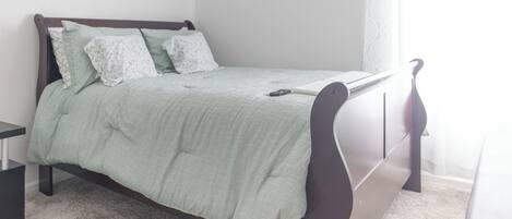 A neatly made bed with a green bedspread and white pillows, a bedside table, and a window with sheer curtains.