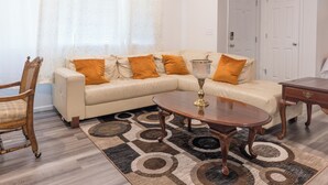 Ein Wohnzimmer mit einem beigen Sofa, zwei orangefarbenen Kissen, einem Holztisch und einem gemusterten Teppich.