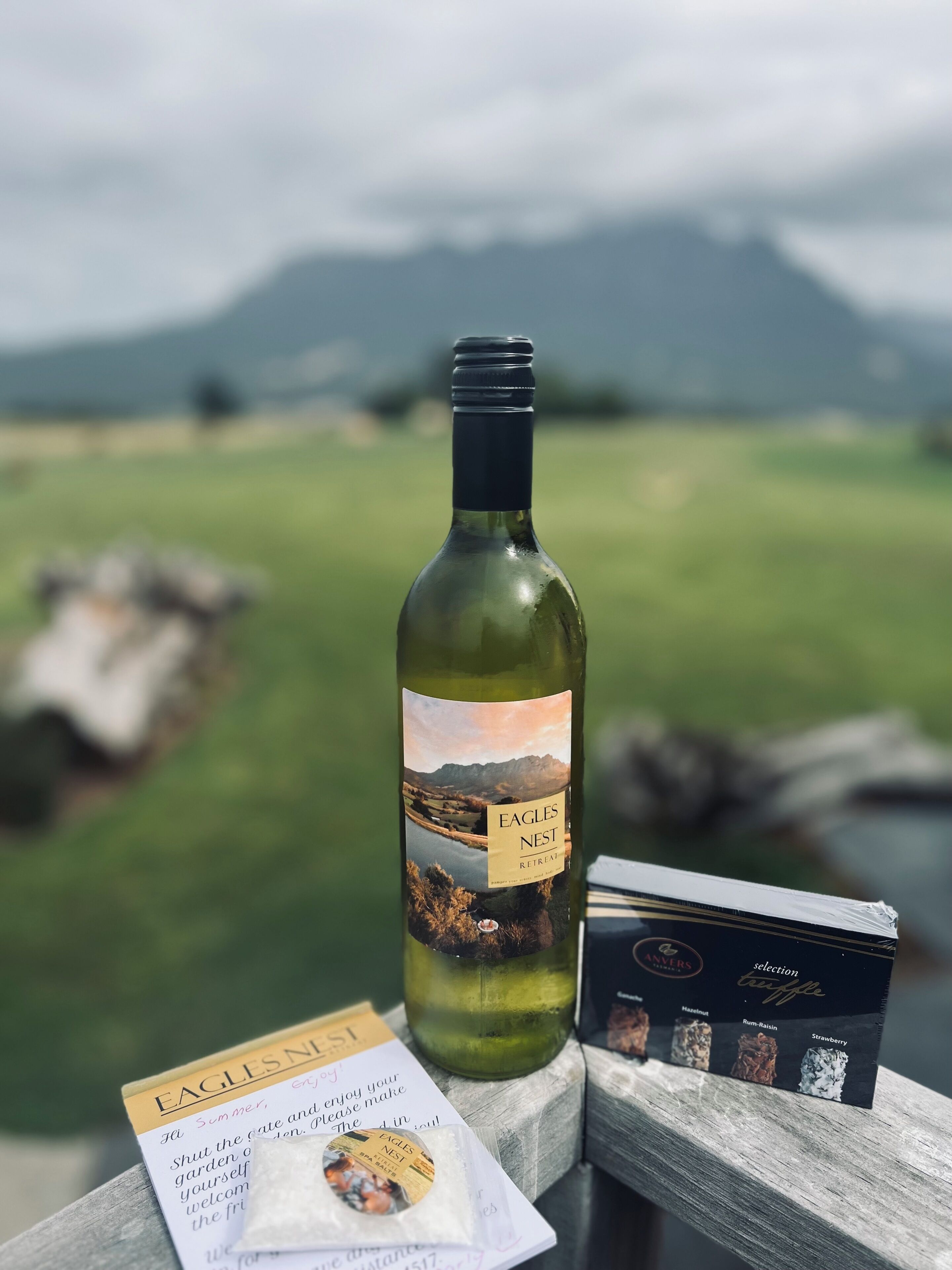 Una bottiglia di vino, una scatola di cioccolatini e un biglietto su un piano di legno con vista panoramica sullo sfondo.