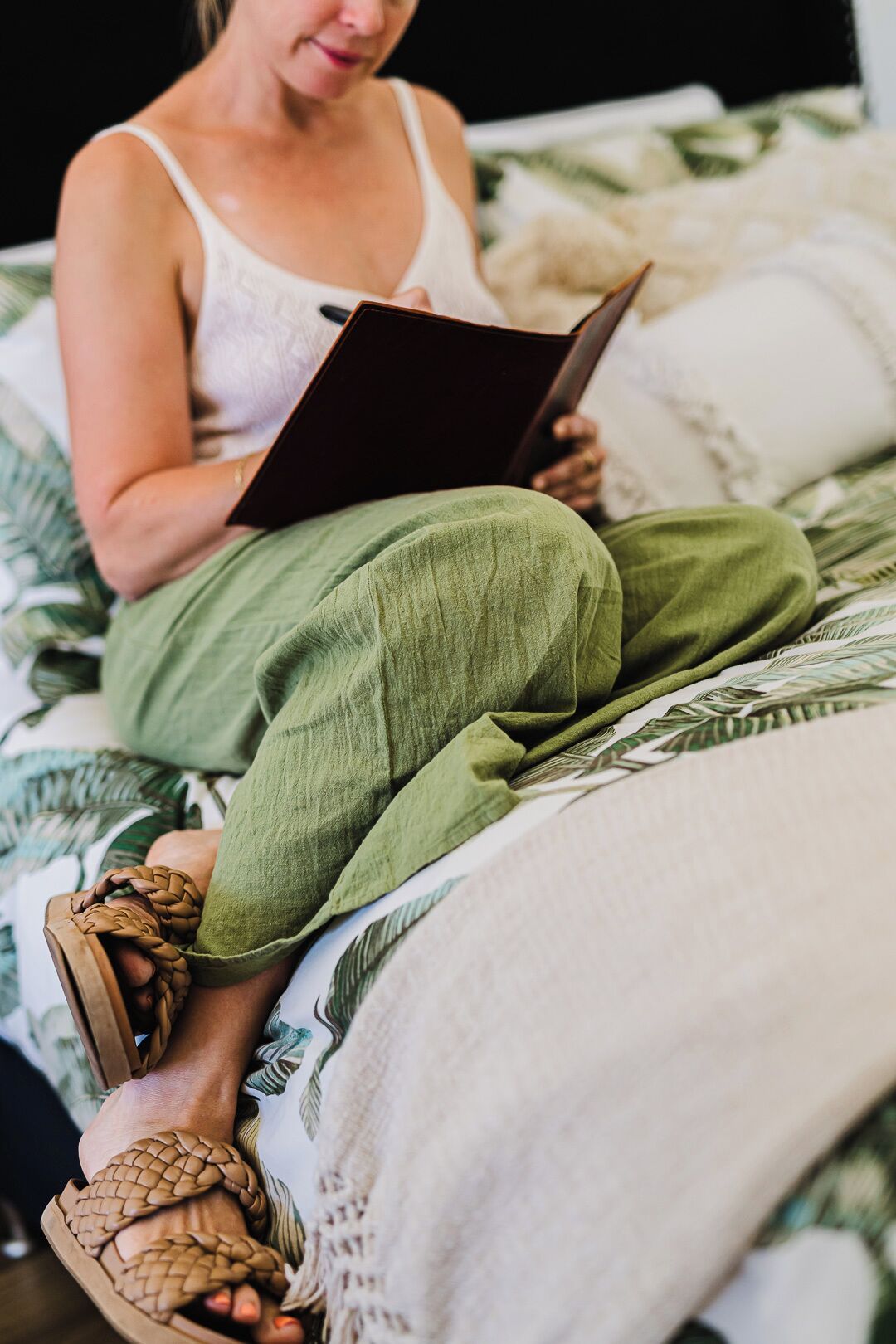 Una persona sentada en una cama con colcha estampada, vestida con una camiseta blanca y pantalones verdes, sosteniendo un libro.