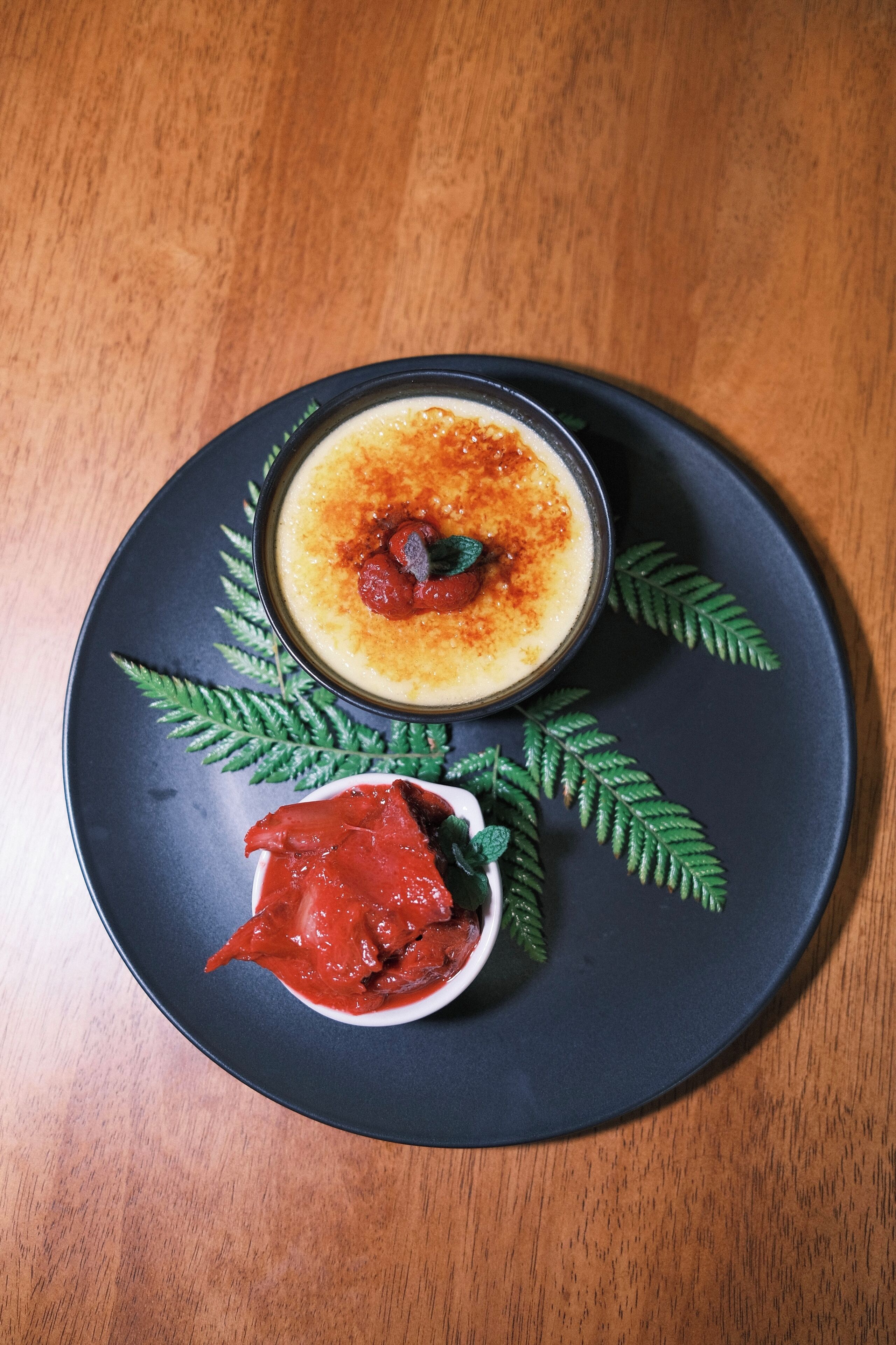 En desserttallrik med en krämig rätt, en liten skål med röd sås och dekorativa ormbunksblad.