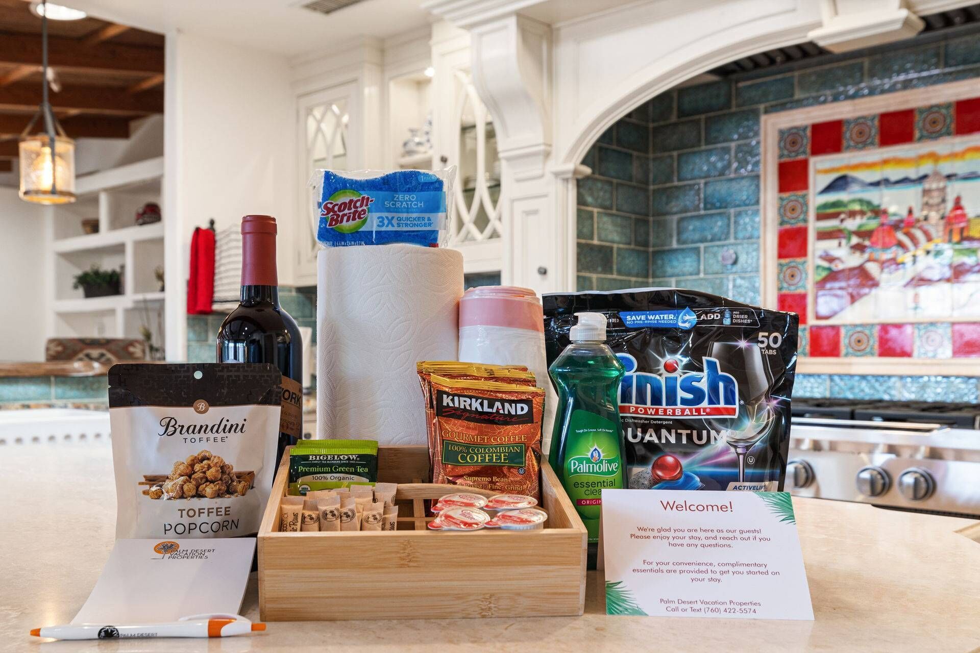 A kitchen counter with various food and drink items, including a bottle of wine, a box of Scotch-Argie Zero Scratch, a pack of Kirkland Signature Gourmet Coffee, a box of Bigelow Premium Green Tea, a box of Palmolive Essential Clean, a pack of Finish Powerball detergent, and a Brandini Toffee Popcorn box.