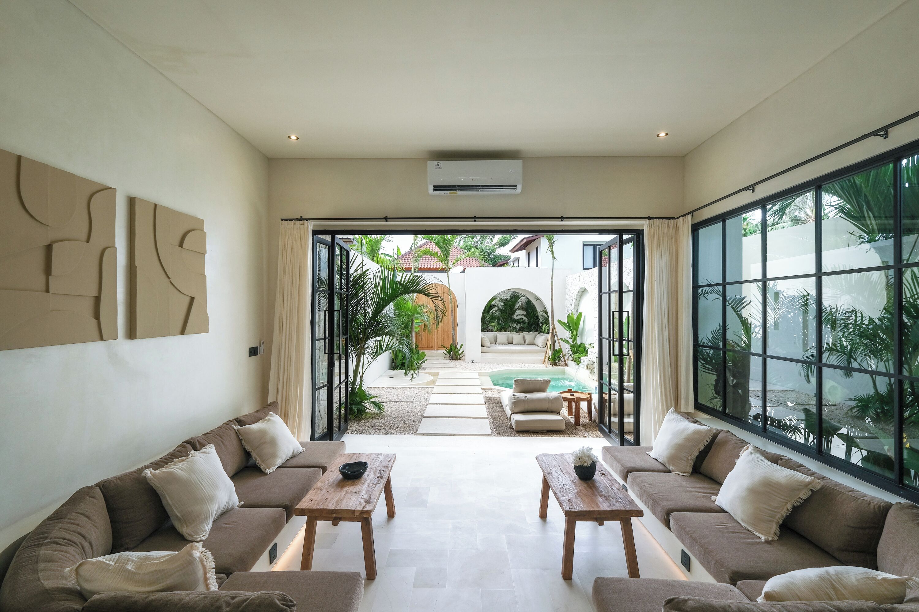 Ein modernes Wohnzimmer mit einer Sofagarnitur, zwei Holznachttischen und einem großen Fenster mit Blick auf einen Pool und eine Außenanlage.