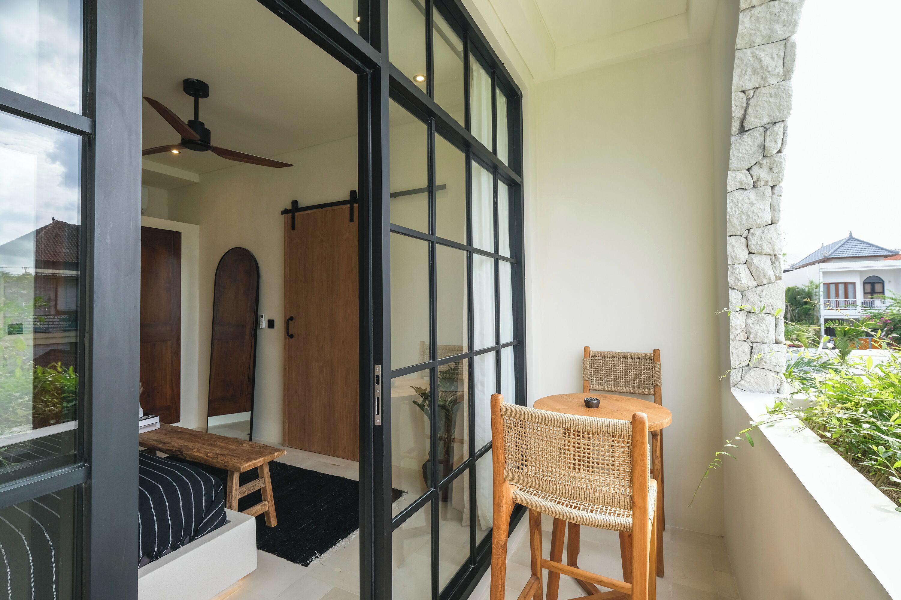 A balcony with a wooden table and chairs, a bench, and a view of a house through large glass doors.