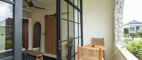 A balcony with a wooden table and chairs, a bench, and a view of a house through large glass doors.