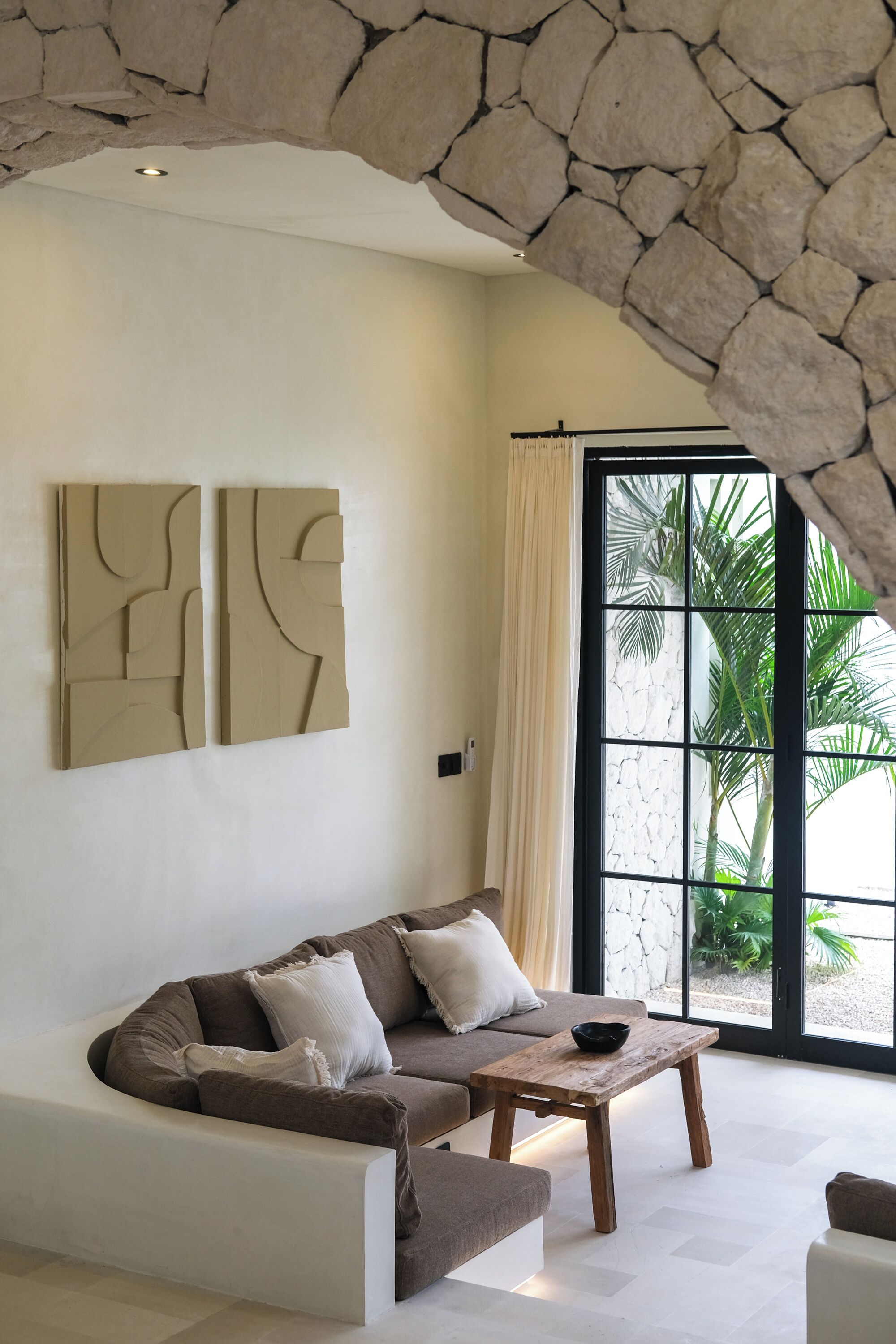 A modern living room with a stone arch ceiling, a large window with a view of palm trees, a brown sectional sofa, and abstract wall art.
