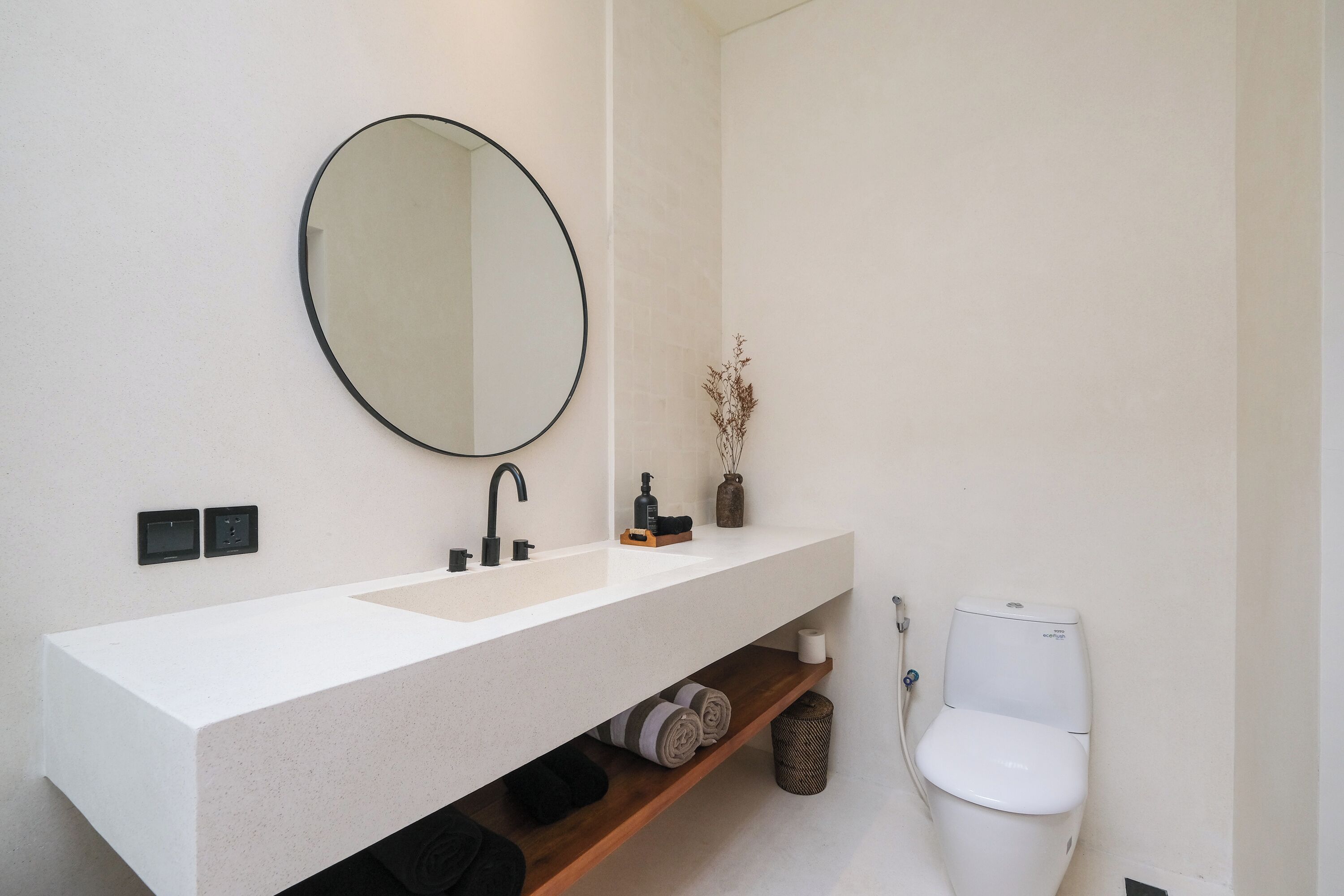 Une salle de bain moderne avec un lavabo blanc, un robinet noir et un miroir rond. On trouve des toilettes à côté du lavabo, et une étagère en dessous, où sont rangées des serviettes et un panier.