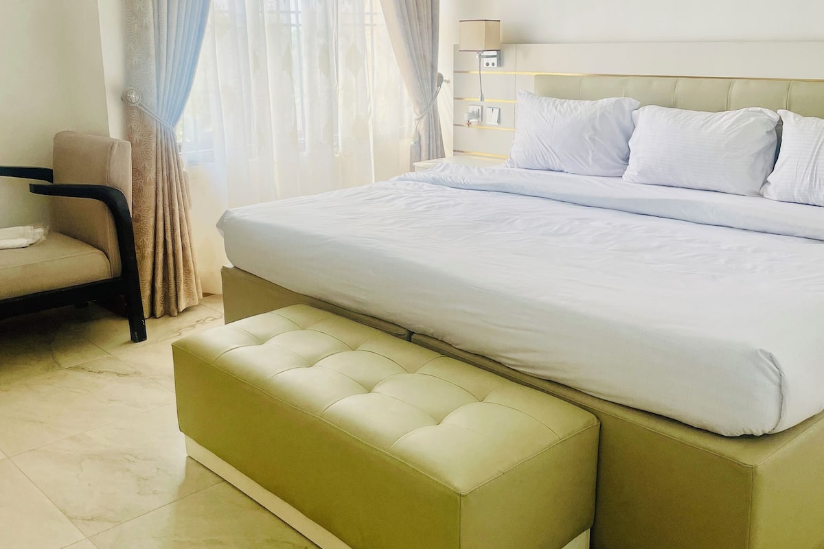 Une chambre d’hôtel avec un grand lit, une table de chevet avec une lampe, une chaise et une fenêtre avec des rideaux.