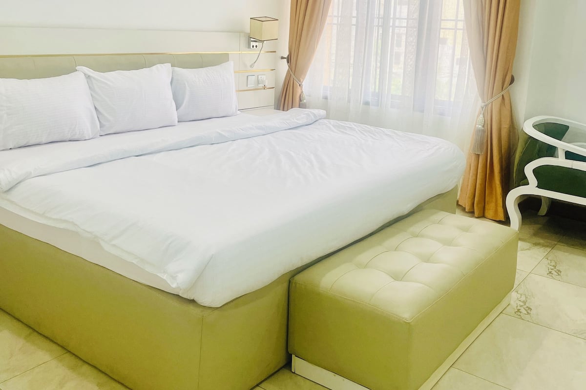 Un lit bien fait, avec du linge de lit blanc, un banc recouvert d’un tissu vert et une petite table accompagnée d’une chaise verte.