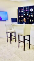 Zona de recepción con dos sillones blancos acolchados, un televisor de pantalla plana en la pared y una estantería con varias botellas.