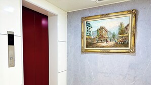 A hallway with a marble wall, a red elevator door, and a framed painting of a historical scene. - ONE STYLE (Naha)