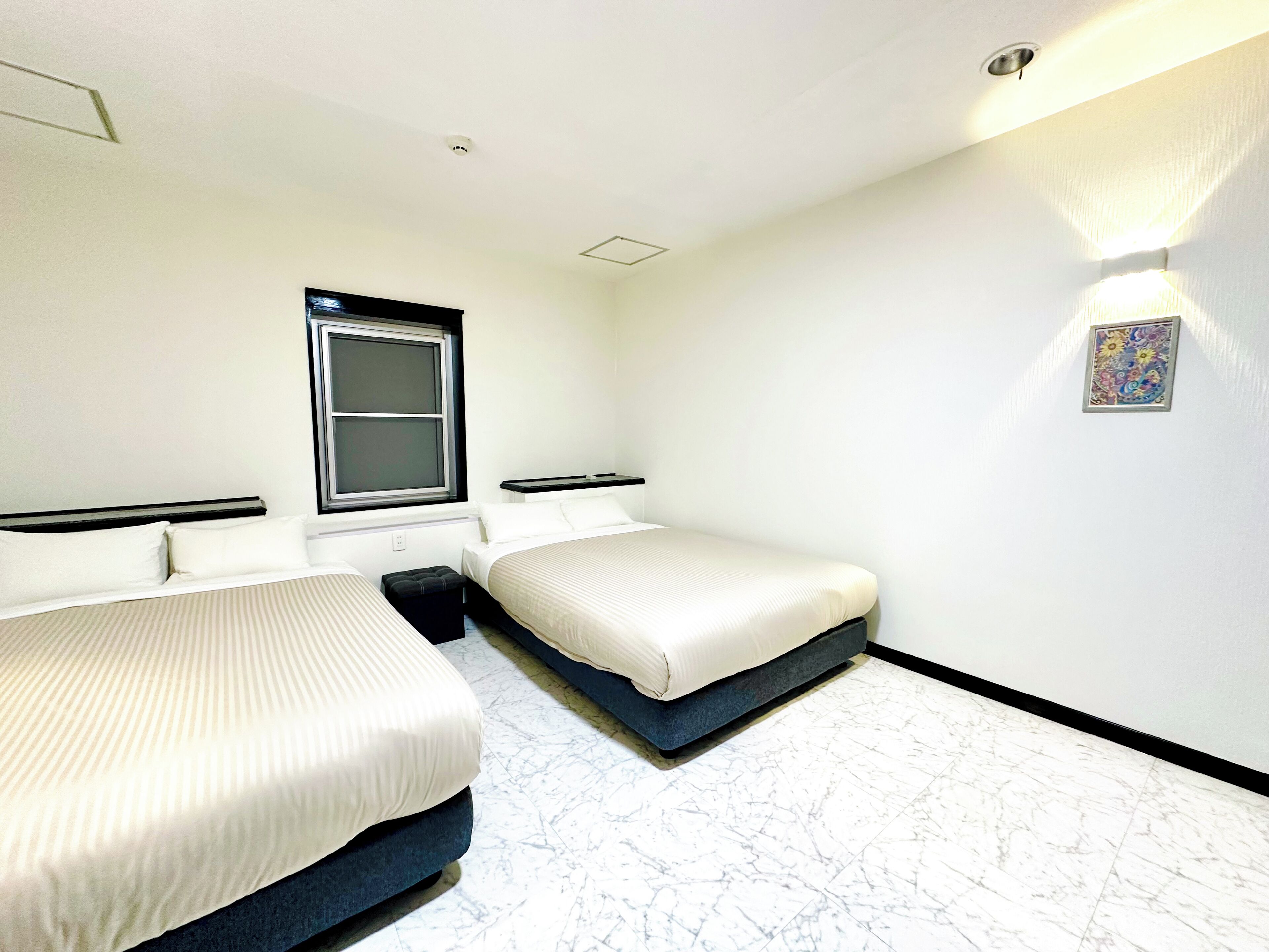 A hotel room with two beds, a window, a wall-mounted light, and a marble floor.