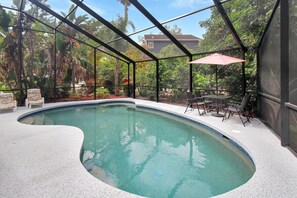 Une piscine à bords incurvés, entourée d’un patio couvert avec des chaises et un parasol.