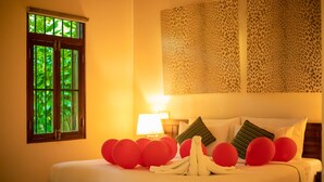 A hotel room with a bed, red balloons, a window with greenery, and a patterned wall.