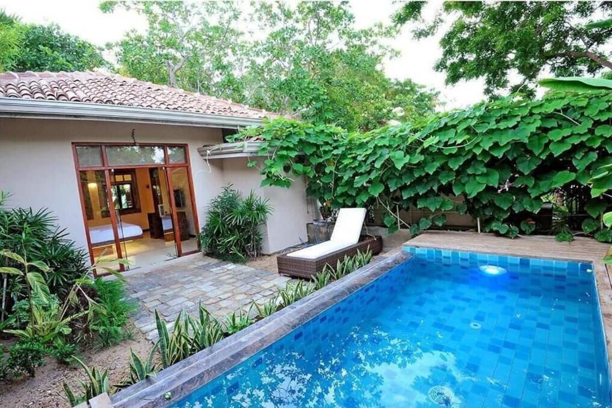 A swimming pool with a tiled edge, a lounge chair, and a building with a tiled roof and glass doors.