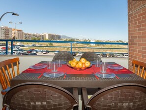 A table set for two with a red tablecloth, plates, cutlery, and a bowl of fruit. The table is outdoors with a view of a parking lot and buildings in the background.