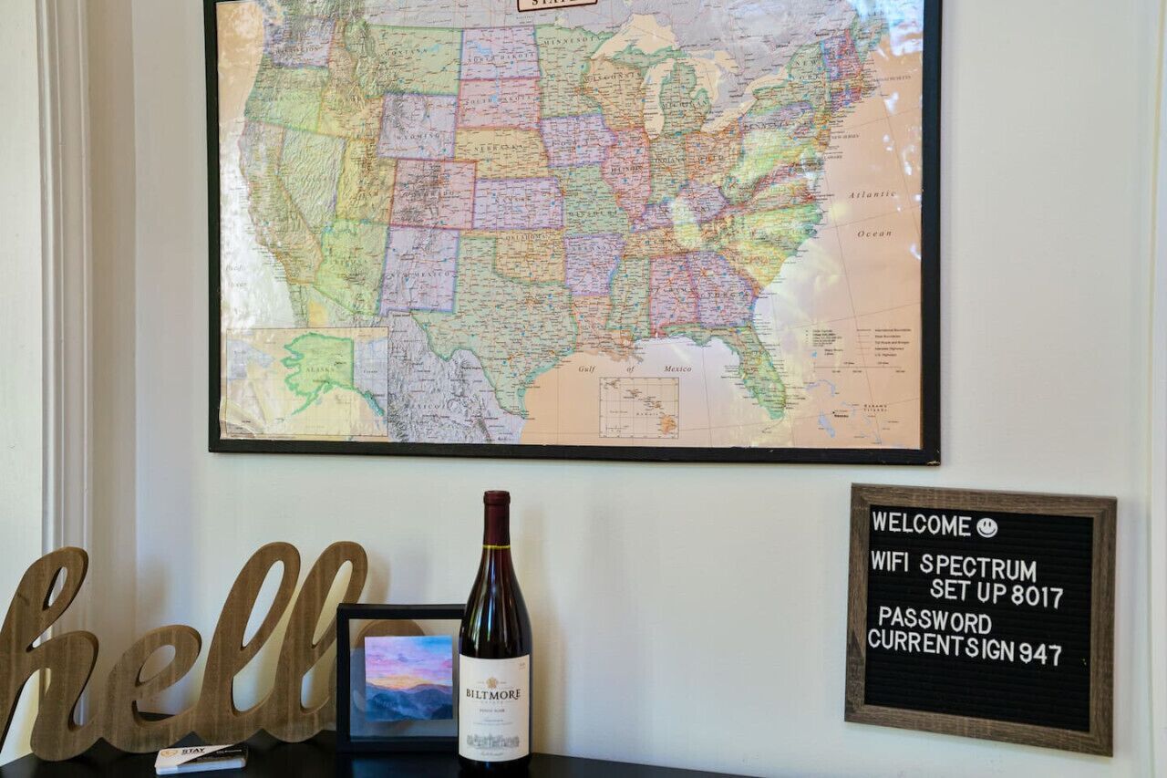 A wall with a large map, a framed welcome sign, a bottle of wine, and a 'hello' sign.
