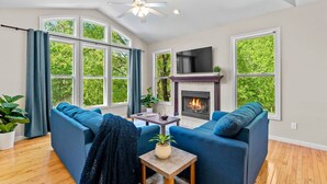 A modern living room with a fireplace, two blue sofas, a ceiling fan, and large windows offering a view of greenery. - Theatre~2 Hottubs~MTN Views~Hidden SpeakEasy (Swannanoa)