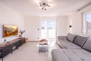 A modern living room with a grey sofa, a coffee table with books and a tray of muffins, a painting of a landscape, and a ceiling light fixture. - City Seaview Apartment Vigo (Rijeka)