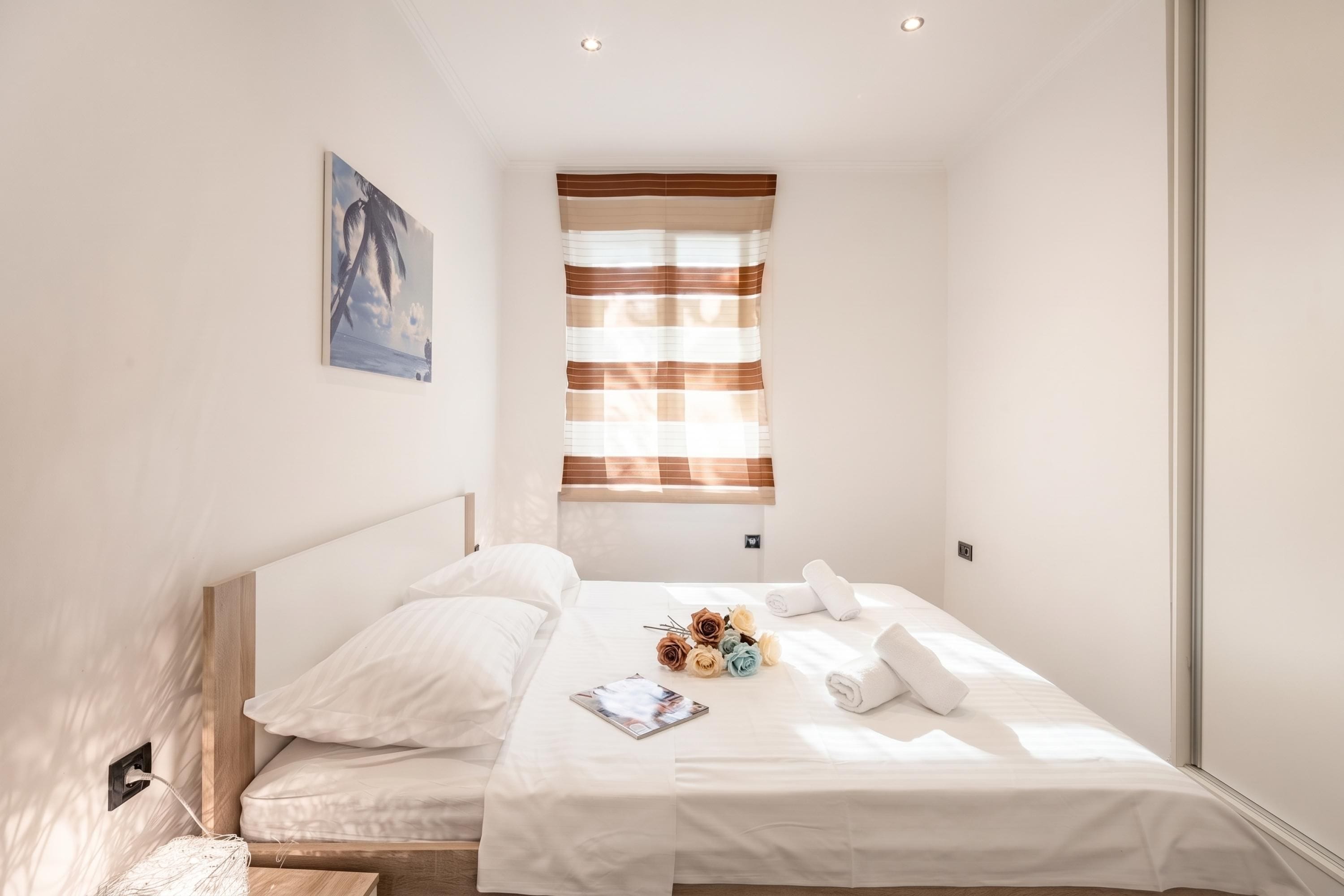 A hotel room with a bed, a window with blinds, a painting on the wall, and a power outlet.