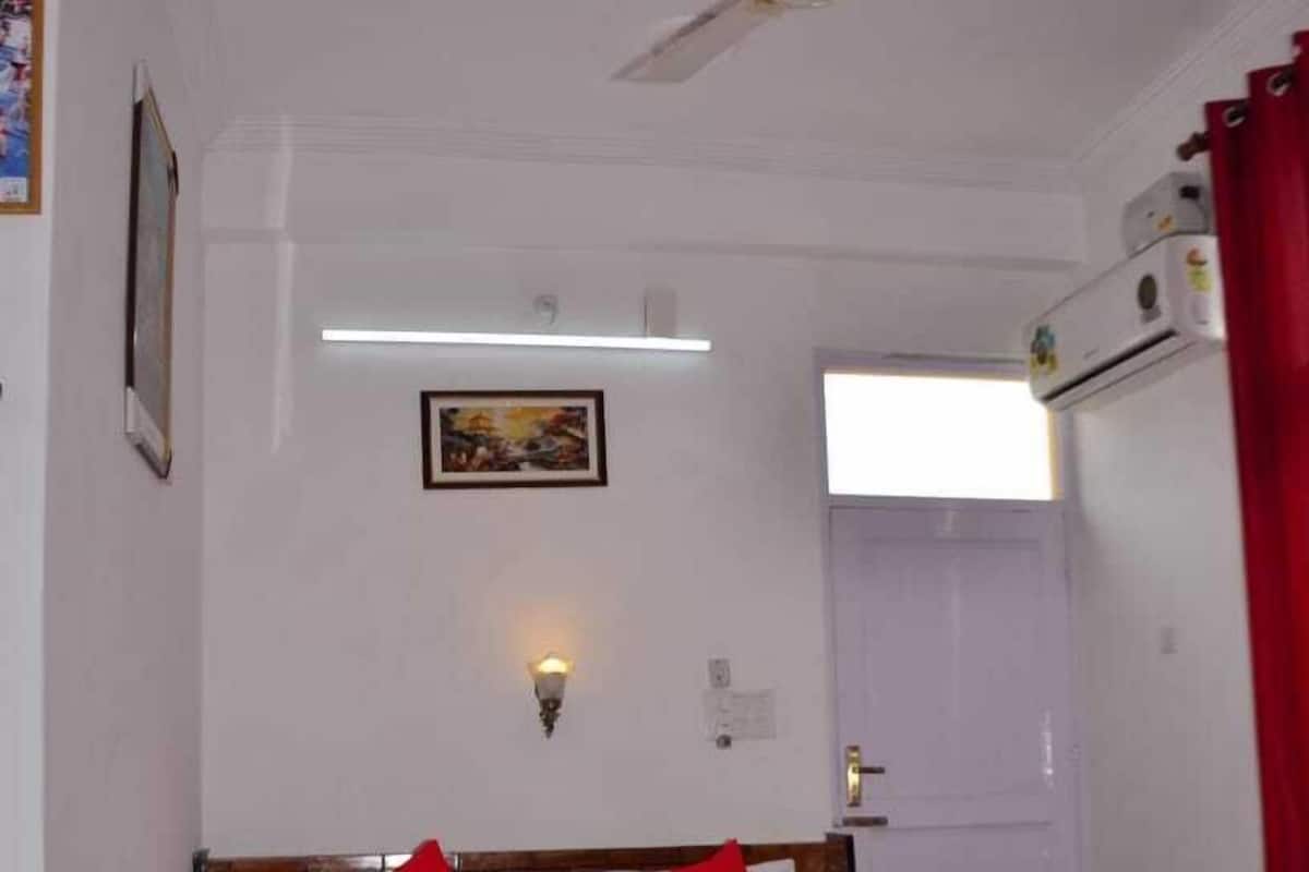 A bedroom with a wooden headboard, a bed with red and white bedding, a ceiling fan, a wall-mounted light, and an air conditioning unit.