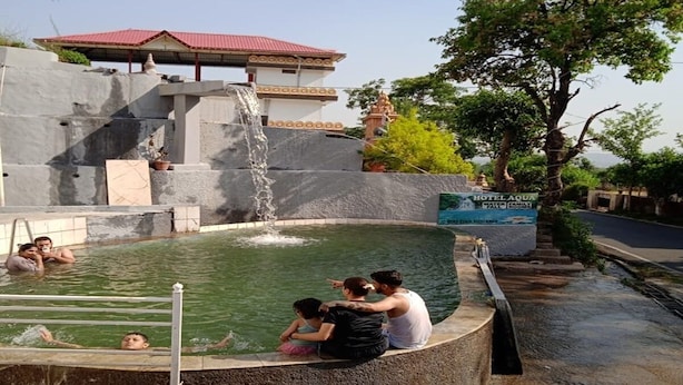 Una piscina con cascada, rodeada por un muro de concreto y un edificio con techo rojo.