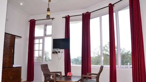 Una habitación con chimenea de ladrillo, televisor montado en la pared, mesa de madera con sillas y ventanas amplias con cortinas rojas.