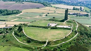 Eine Vogelperspektive auf eine ländliche Landschaft mit einem markanten Turm, einem großen offenen Feld und einem kleinen Gebäude.