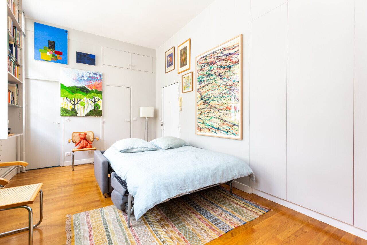 A bedroom with a bed, a chair, a desk, a bookshelf, and artwork on the walls.