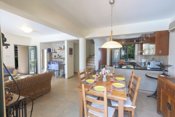 A dining area with a wooden table set for six, surrounded by chairs. A kitchen with modern appliances is visible in the background.