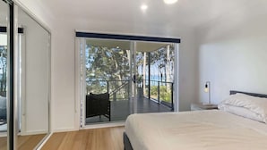 A bedroom with a bed, a nightstand, a lamp, and a balcony with a view of trees and the sea. - Pet-Friendly 3-Bed Coastal Gem Near Long Beach (Long Beach)