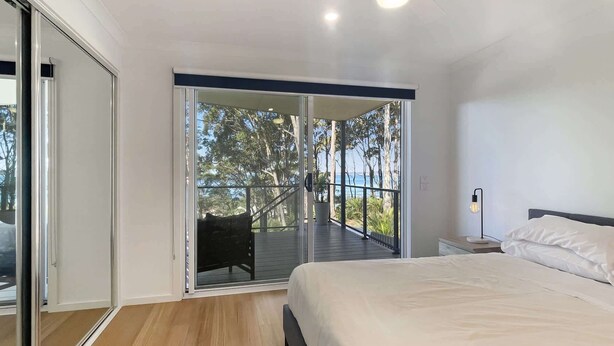 A bedroom with a bed, a nightstand, a lamp, and a balcony with a view of trees and the sea.