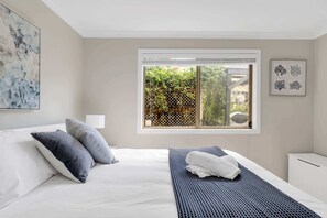 Un dormitorio con cama, almohadas, una mesita de noche, una ventana que da a zonas verdes y un cuadro en la pared.