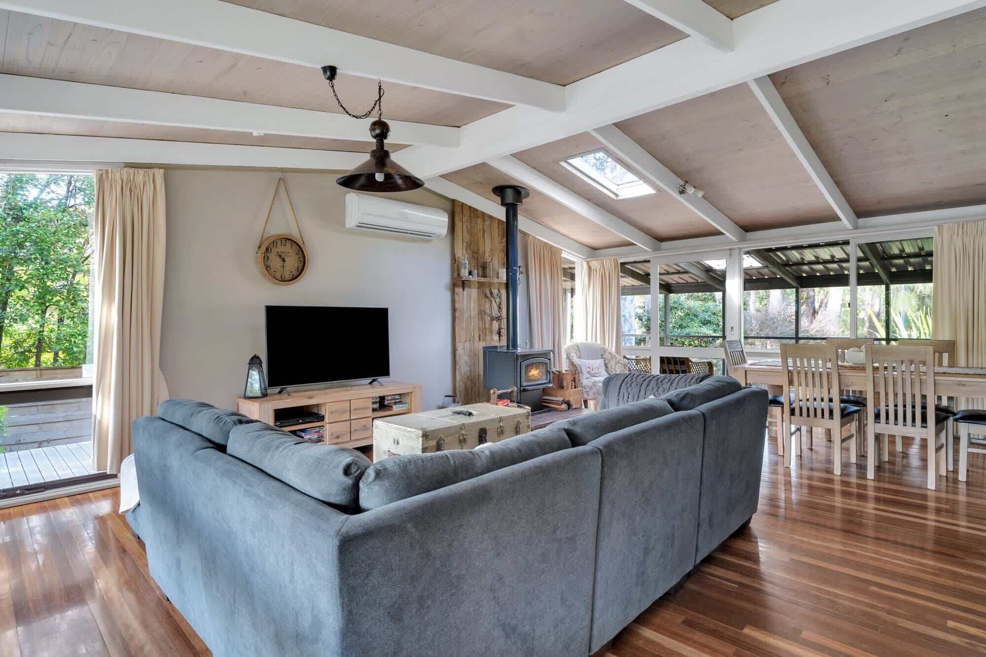 A spacious living room with a large grey sofa, a flat-screen TV, a wooden table, and a built-in wood stove.