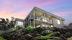 Une maison moderne à deux étages, dotée d’un balcon et entourée de grands rochers et de verdure.