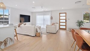 Een moderne woonkamer met een bank, een kleinere zithoek, een eettafel met stoelen, een aan de muur bevestigde televisie en een groot raam met gordijnen.