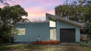 A modern single-story house with a flat roof, a garage with a roller door, and a small front porch with steps. - Cosy Getaway with Fireplace and Spacious Backyard (Broulee)