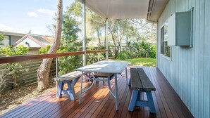 A covered outdoor area with two picnic tables, a wooden deck, and a view of a backyard with trees and a fence. - Cosy Getaway with Fireplace and Spacious Backyard (Broulee)